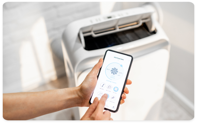 This image focuses on a user’s hand holding a smartphone to control a sleek, portable air filtration or ventilation unit. The mobile app interface displays real-time data on air quality and fan speed, allowing for remote climate management. This represents the "smart office" or workspace trend, where IoT (Internet of Things) devices ensure a personalized and healthy environment through mobile connectivity.